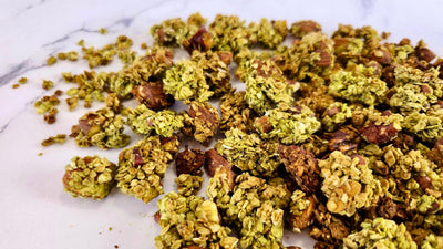 Granola maison au thé matcha, d'un vert tendre et en gros clusters avec des noix de pécan, étalé sur un plan de travail en marbre