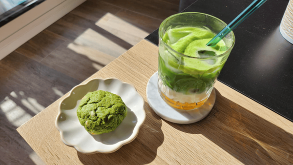 Cookie au matcha vert sur une petite assiette en forme de fleur, à côté d'un verre de matcha latté glacé, sur une table en bois ensoleillée