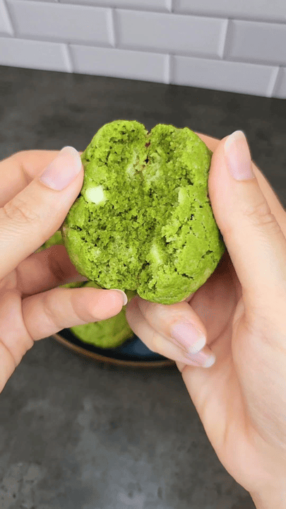 Cookie au matcha rompu en deux entre deux mains, révélant un cœur vert moelleux parsemé de pépites de chocolat blanc