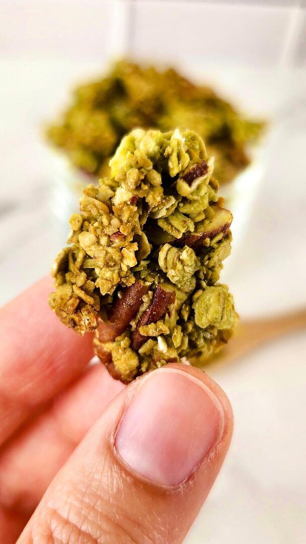Gros plan d'une grosse pépite de granola au matcha tenue entre les doigts, avec amandes et noix de pécan visibles dans le cluster vert