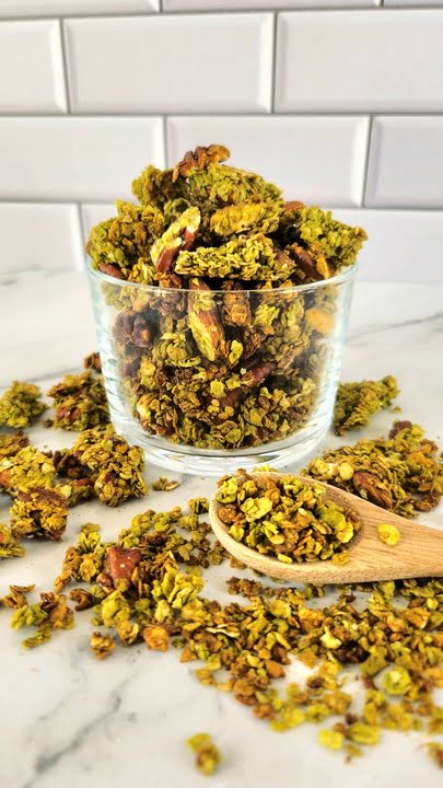 Bol en verre débordant de granola maison au matcha, d'un vert tendre, avec une cuillère en bois sur un plan de travail en marbre
