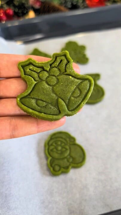 Sablé de Noël au matcha en forme de cloche, aux motifs nettement embossés, tenu à la main au-dessus de la pâte découpée