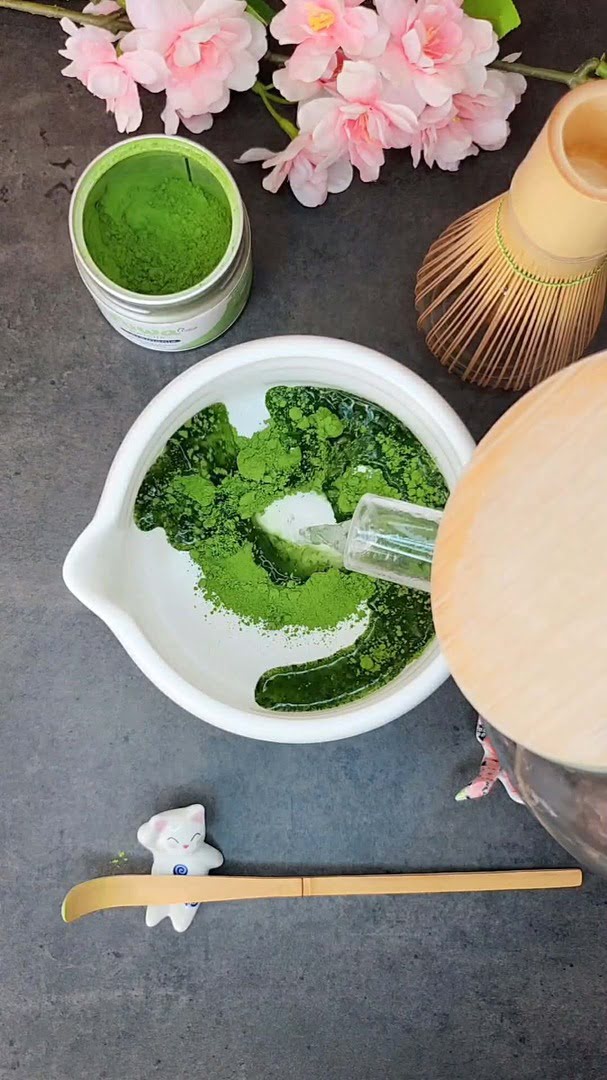 Eau versée sur la poudre de matcha dans un bol blanc, à côté d'un chasen en bambou et de fleurs de cerisier